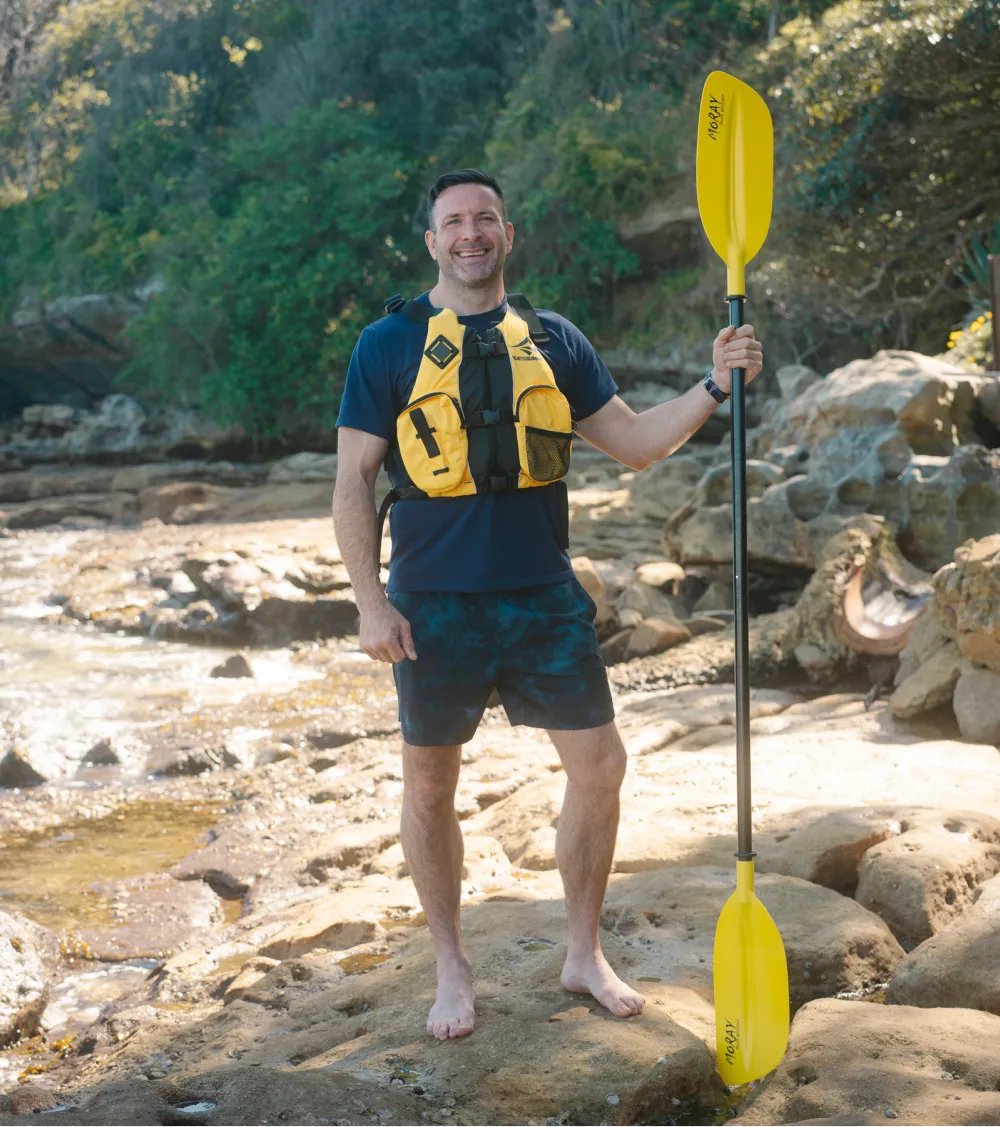 Founder Chris of Sydney Kayaking Tours smiling and holding a yellow paddle on a rocky beach